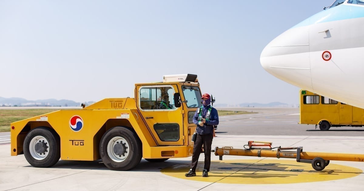 Ground agent standing beside a tug with towbar attached to the nose of a 737, with a baggage truck visible in the background