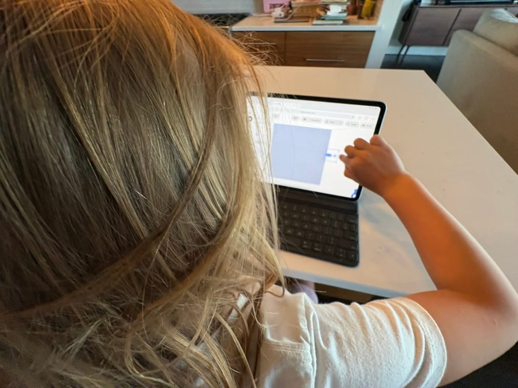 A young child at a kitchen table reaching out to drag an aircraft on the AirPlx base view, shown on an iPad propped in a keyboard case