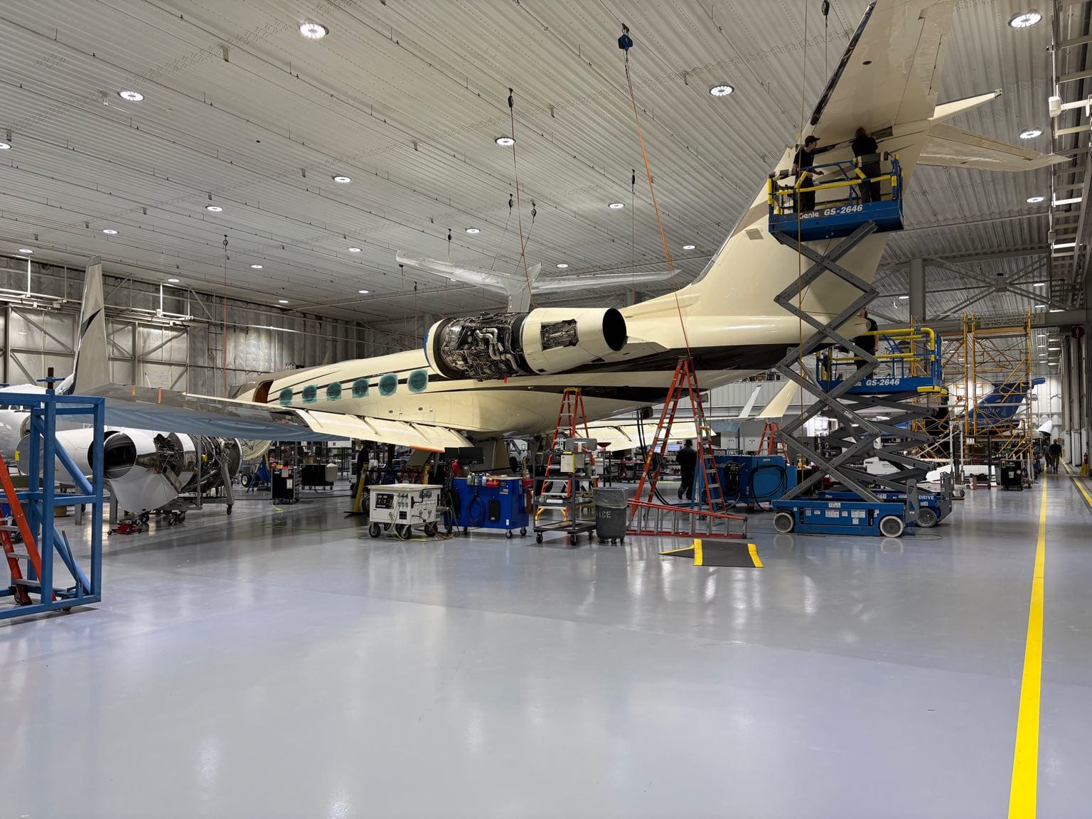 Two technicians on a scissor lift working the tail of a business jet in a Duncan Aviation hangar, its engine open and a second aircraft under maintenance alongside it