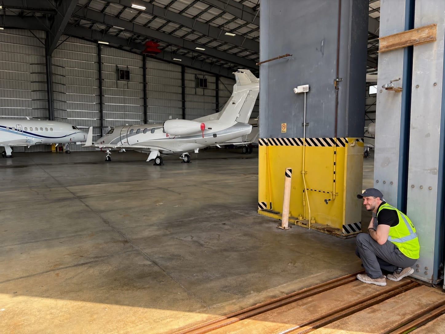An AirPlx team member crouched at a hangar door jamb measuring the opening, with two business jets parked inside the hangar behind him