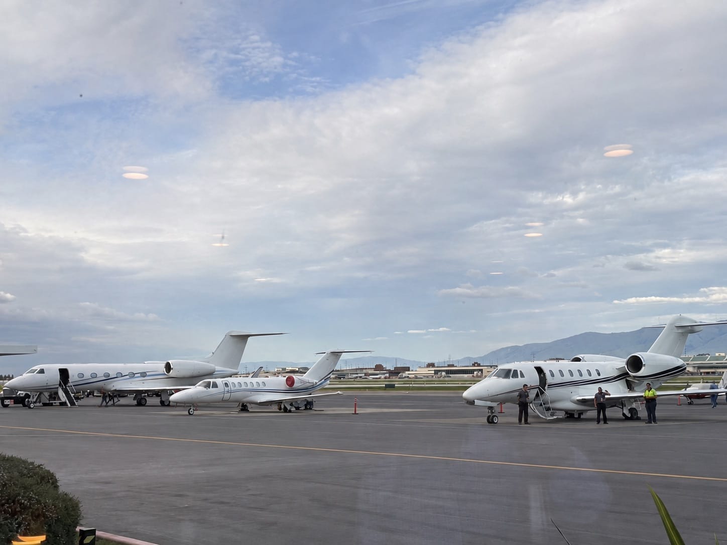 Business jets parked close together on a ramp in the evening, with line crew working between them, hangars and airport buildings behind, and mountains on the horizon